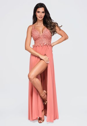 Femme en robe rose corail avec corsage en dentelle florale et fente haute, posant avec une main sur la hanche et une jambe partiellement exposée.