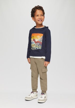 Marinefarbener Hoodie mit langen Ärmeln und Grafikdruck, helles Shirt darunter, beigefarbene Cargo-Hosen und weiße Sneaker mit grauen Akzenten.