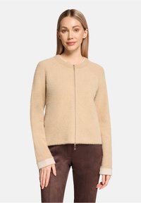 Pull beige à fermeture éclair fabriqué en tissu doux et texturé, avec un col rond et des poignets côtelés contrastants. Porté sur un pantalon marron.