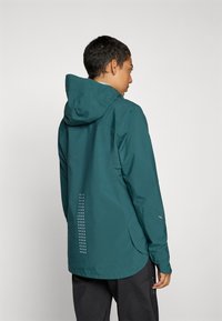 Jachetă impermeabilă de culoare teal, cu glugă, având un design reflexiv punctat pe spate și un hem ușor curbat. Textură moale și design minimalist.