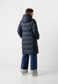 Marineblå pufferjakke med hette, med en quiltet design, lange ermer og midt-til-lår lengde. Kombinert med vide denimbukser.