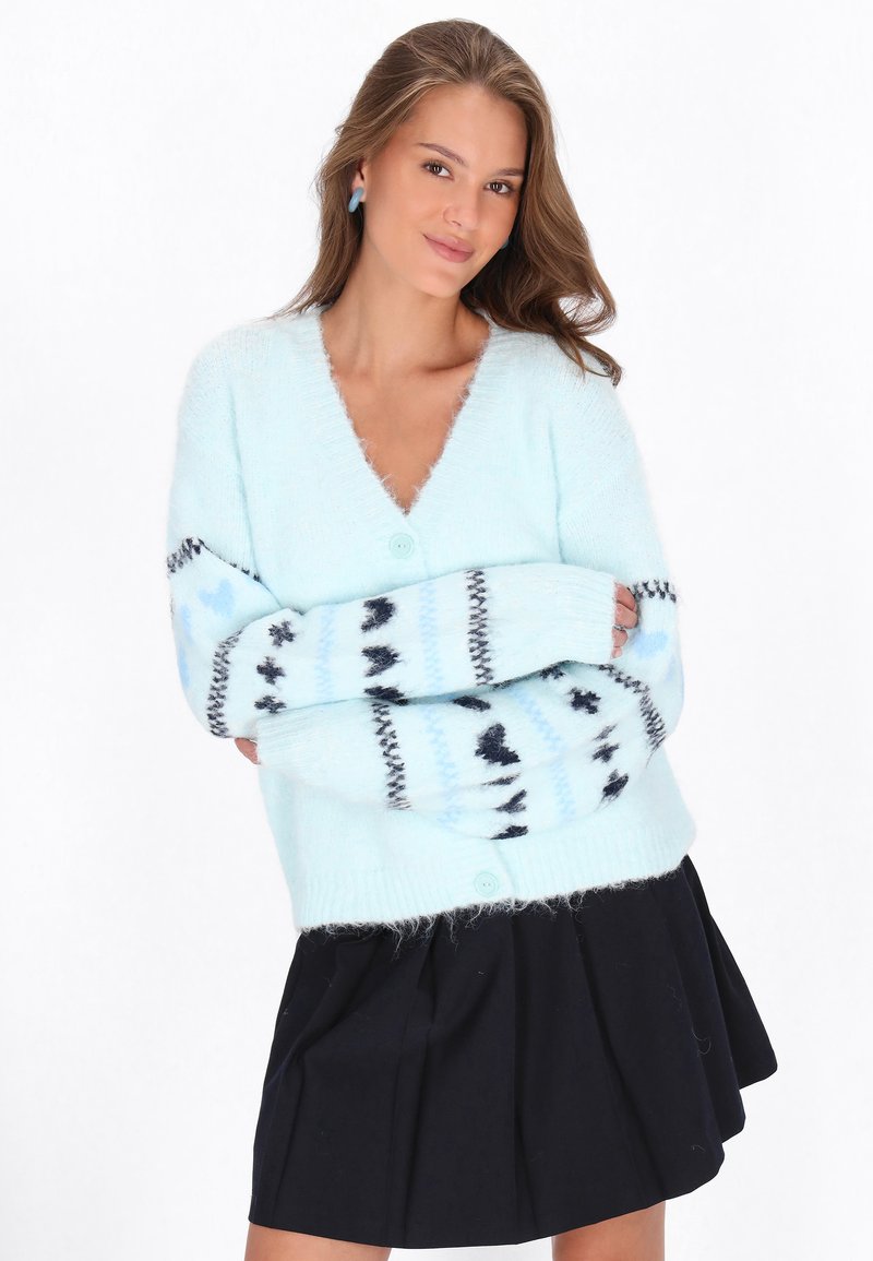Ljusblå fluffig cardigan med V-ringning, med mönstrade ärmar i mörkare blått och svart. Matchad med en svart plisserad kjol.