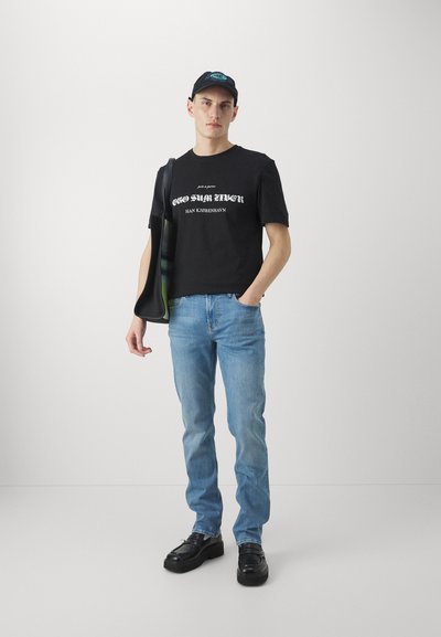 Schwarzes Baumwoll-T-Shirt mit weißem Text, hellblaue Jeans und schwarze Schuhe. Das Modell trägt zudem eine schwarze Mütze und eine Umhängetasche.