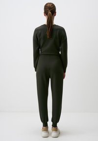 Dunkelgrüner Sweatshirt und passende Jogginghose, aus weichem Stoff, mit gerippten Bündchen und einem elastischen Bund. Weiße Sneaker sind sichtbar.