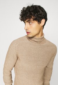 Pull à col roulé beige en côtes avec des manches longues, présentant un motif de tricot texturé et une silhouette ajustée. Matière douce avec une finition extensible.