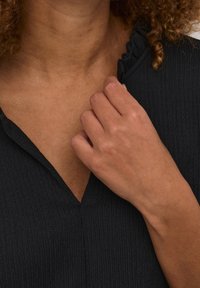 Sort tekstureret top med V-udskæring og flagrende detaljer ved kraven, hvor en hånd forsigtigt griber fat i stoffet nær udskæringen.