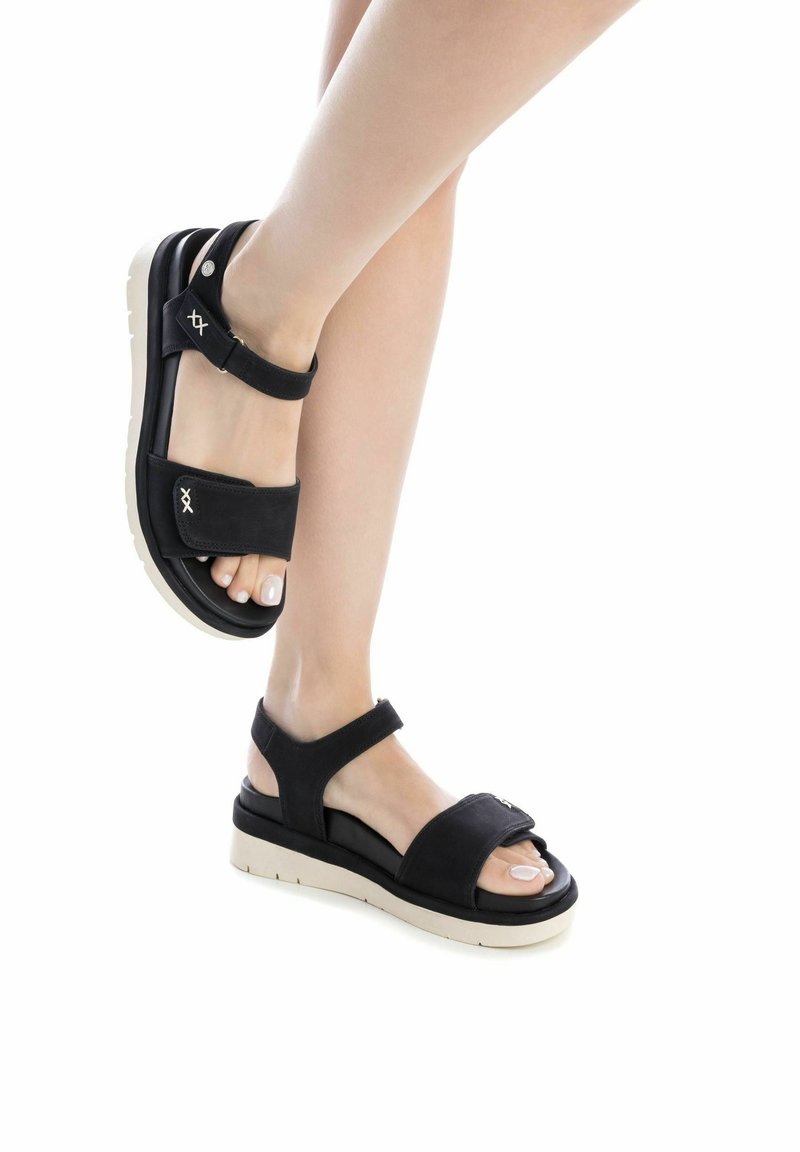 Sandalias negras con dos correas ajustables, una plantilla acolchada y suela de color claro. Presenta un detalle decorativo en la correa.
