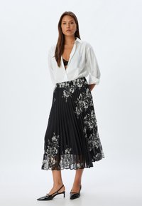 Jupe plissée noire avec motif floral, chemise blanche à boutons, et escarpins à talons ouverts noirs. Tissu lisse et texture fluide.