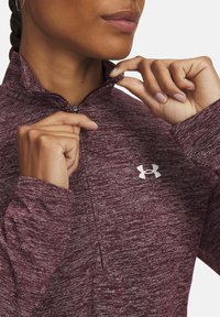 Mørkerød zip-up atletisk top med tekstureret stof, der har en høj krave og logo accent på brystet, hænder der griber fat om kraven.