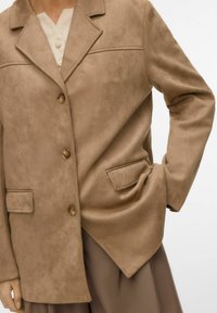 Gros plan sur une personne portant un blazer en daim couleur tan avec trois boutons, des poches à rabat, et un haut beige en dessous, la main dans la poche.