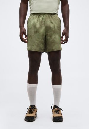 Mand iført olivengrønne shorts, beige sokker og beige sorte sneakers stående foran en ensartet baggrund.