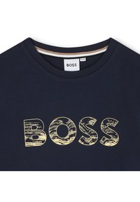 Tamsiai mėlyna medvilninė marškinėliai su apvalia iškirpte, su tekstūruotu geltonu "BOSS" logotipu bangų raštu. Prie krūtinės matoma etiketė.