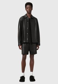 Schwarze Lederjacke mit Druckknöpfen, schwarzes Textilshirt und passende Shorts. Das Model trägt schwarze Schuhe mit weißen Socken. Minimalistisches Design.