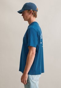 Niebieski bawełniany t-shirt z krótkimi rękawami, o luźnym kroju, z grafiką na plecach. Połączony z jasnoniebieskimi dżinsami i niebieską czapką.