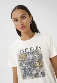 T-shirt en coton crème avec un graphique floral comprenant des fleurs bleues et jaunes, entourées de noir, et le texte "LES FLEURS" au-dessus du design.
