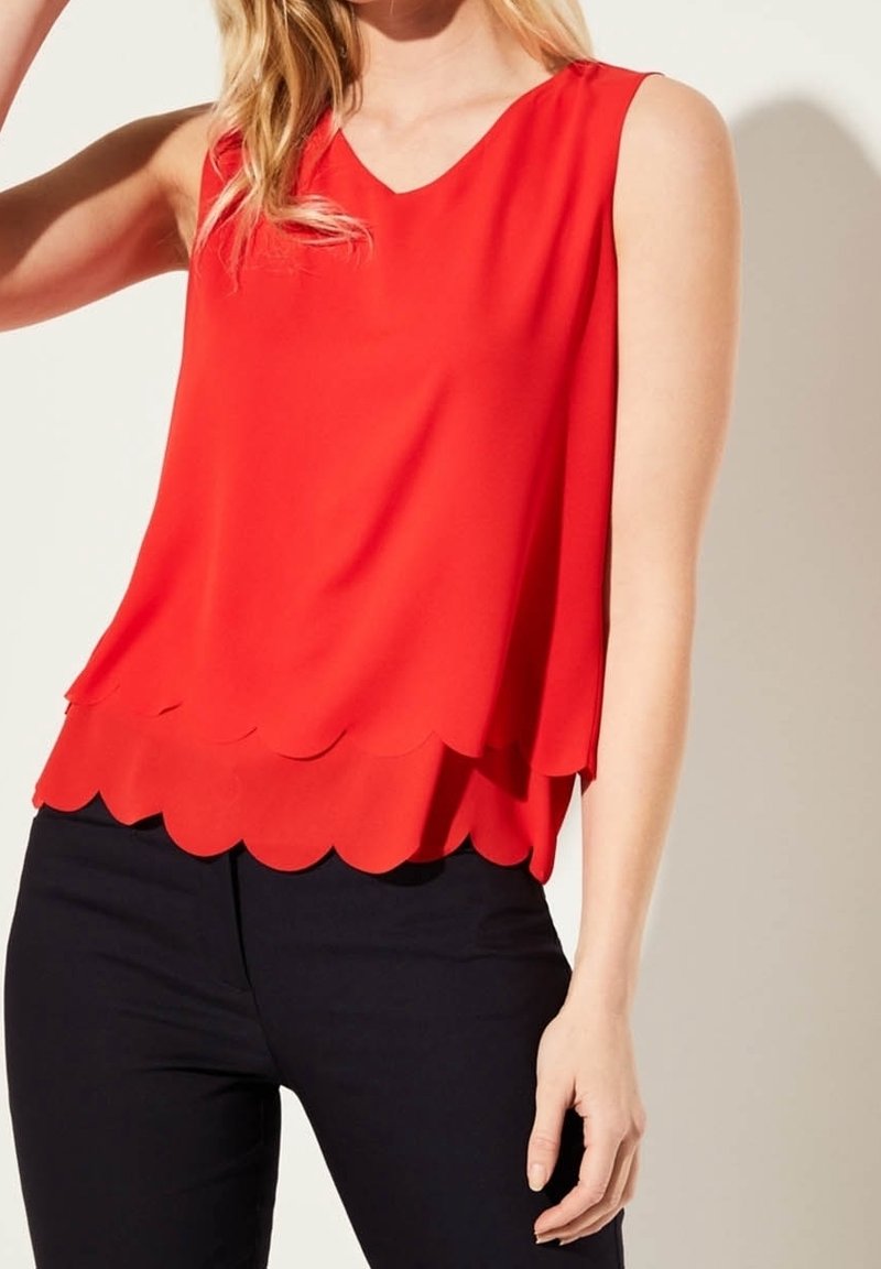 Femme portant un haut rouge sans manches avec un ourlet double couche festonné et un pantalon noir, debout devant un fond clair uni.