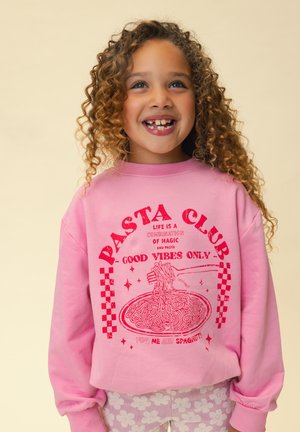 Lachend kind met krullend haar draagt een roze sweatshirt met de tekst "Pasta Club, alleen goede vibes" en een illustratie van een vork die spaghetti oprolt.