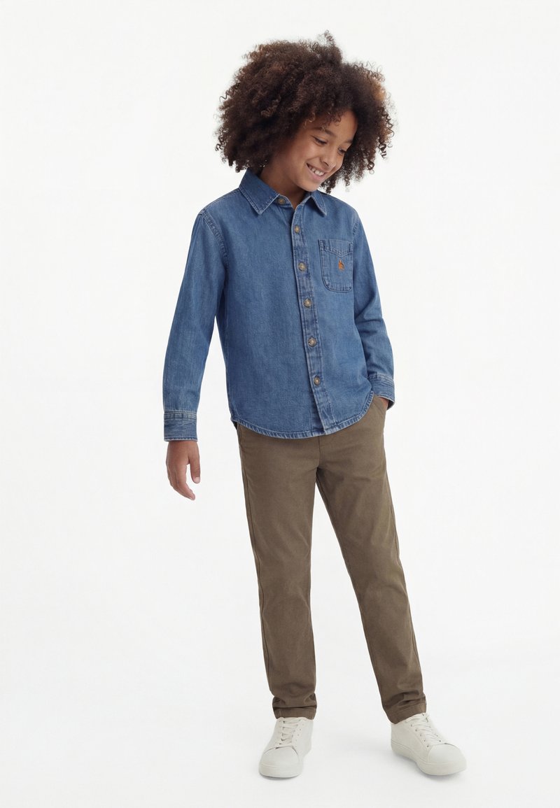 Usmievajúce sa dieťa kučeravými vlasmi, oblečené v modrej rifľovej košeli, hnedej nohaviciach a bielych teniskách, stojace s jednou rukou v vrecku.