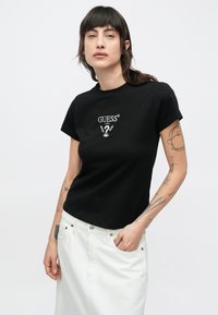 Μαύρο βαμβακερό μπλουζάκι με στρογγυλή λαιμόκοψη και κοντά μανίκια. Διαθέτει λευκό λογότυπο "GUESS®" και γραφικά στο στήθος.