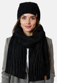 Fekete kötött sál és sapka szett, bordás mintákkal és puha textúrával. A sál egy szürke kabáton lóg, bemutatva a színkombinációt.
