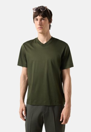 Man draagt een donkergroen V-hals T-shirt, een donkere broek en een zonnebril die op zijn hoofd rust, tegen een effen achtergrond.
