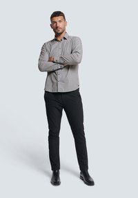 Grijs-witte geruite blouse met lange mouwen, zwarte slim-fit broek en zwarte enkelaarsen, staande pose tegen een lichte achtergrond.