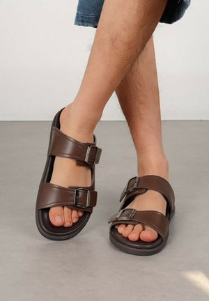 Personne portant des sandales marron foncé à double bride avec des boucles en métal, debout sur un sol gris clair, jambes croisées.