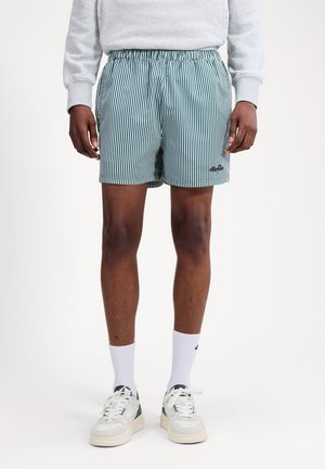 Hombre con pantalones cortos de rayas verticales verdes y blancas, calcetines blancos hasta la mitad de la pierna, zapatillas blancas y sudadera gris claro.