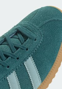 Green suede sneaker with light blue stripes, textured upper, round laces, and a gum rubber sole. Zigzag accents along the stripes.