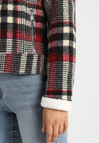 Rød, sort og cremefarvet rudemønstret fleecejakke med en lynlås lomme og hvide sherpa-manchetter. Har blød tekstur og en tætsiddende design.