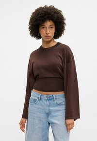 Suéter castanho, cropped, de manga longa em material de malha cintilante, combinado com jeans claros de cintura alta.