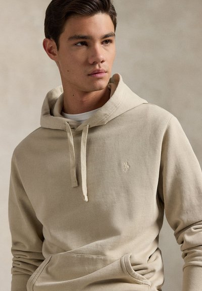 Beige hettegenser med snor, ribbet tekstur, kengurulomme og brodert logo på brystet. Myk stoff med en lett justerbar hette.