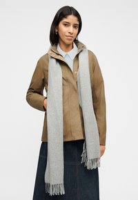 Νεαρή γυναίκα που φοράει μπεζ σακάκι, ανοιχτό μπλε πουκάμισο με γιακά, σκούρα φούστα και μακρύ γκρι κασκόλ με φούντες σε λευκό φόντο.