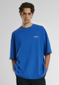 Blå, oversized t-skjorte i bomull med rund hals og korte ermer. Har et lite hvitt brodert "Forbudt"-logo på brystet.