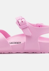 Pink sandal with adjustable buckle, made of textured synthetic material. Open design, flat sole, and logo visible on the insole.