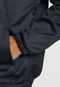 Giacca sportiva blu navy con texture liscia, polsini elasticizzati e tasca laterale; presenta un design minimalista e un tessuto leggero.
