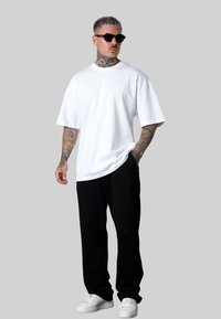 White oversized t-shirt, black sweatpants, white sneakers. Short sleeves, relaxed fit, plain design. Tattoos visible on arms, sunglasses worn.