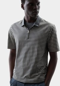 Polo a maniche corte con un motivo geometrico a triangoli in bianco e nero, dotato di colletto con zip e colletto morbido grigio.