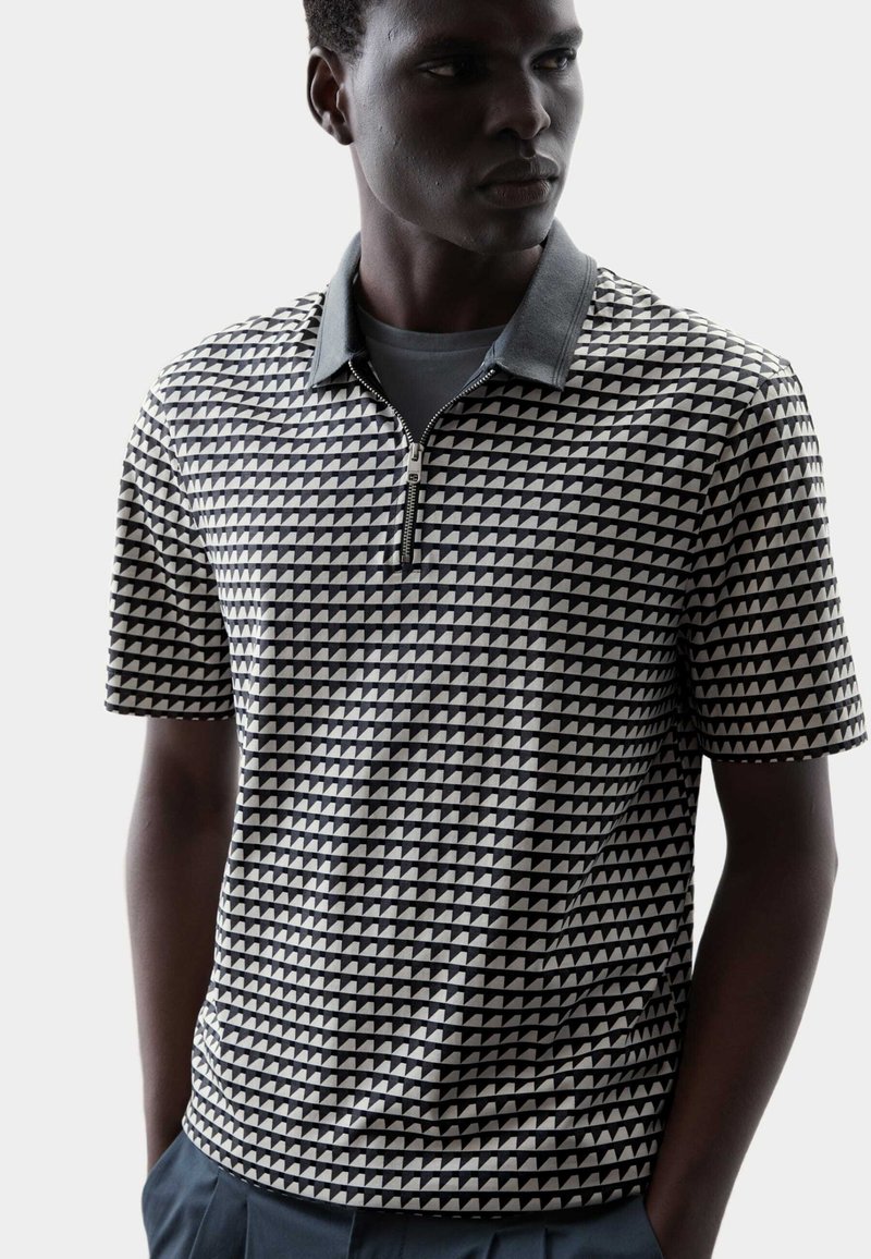 Polo a maniche corte con un motivo geometrico a triangoli in bianco e nero, dotato di colletto con zip e colletto morbido grigio.