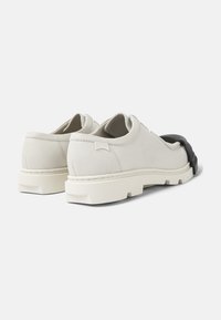 Chaussures en suede gris clair avec un accent noir, présentant une semelle épaisse et un design minimaliste. L'arrière inclut un petit détail de logo.