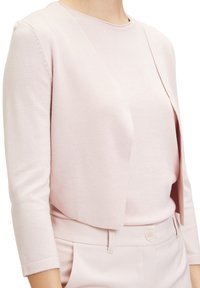 Světle růžový krátký kardigan s měkkým, texturovaným povrchem a otevřeným předním dílem, nošený přes odpovídající top a na míru šité kalhoty.