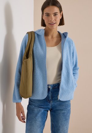 Ung kvinde iført lyseblå fleecejakke og jeans, bærende en olivengrøn skuldertaske, stående mod en beige og hvid væg.