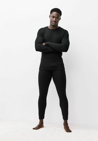 Čierny dlhý rukáv a legíny z rebrovanej tkaniny, s priliehavým strihom a jemnou textúrou, oblečené na mužskej postave s prekríženými rukami.