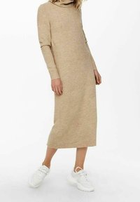 Robe beige en maille à manches longues avec col cheminée et poignets côtelés, s'étendant jusqu'au milieu du mollet. Associée à des baskets blanches épaisses.