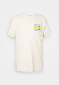 Krémové bavlněné tričko s krátkými rukávy. Na přední straně je textová grafika v modré, žluté a oranžové barvě: "REVOLT VOTE REPEAT".