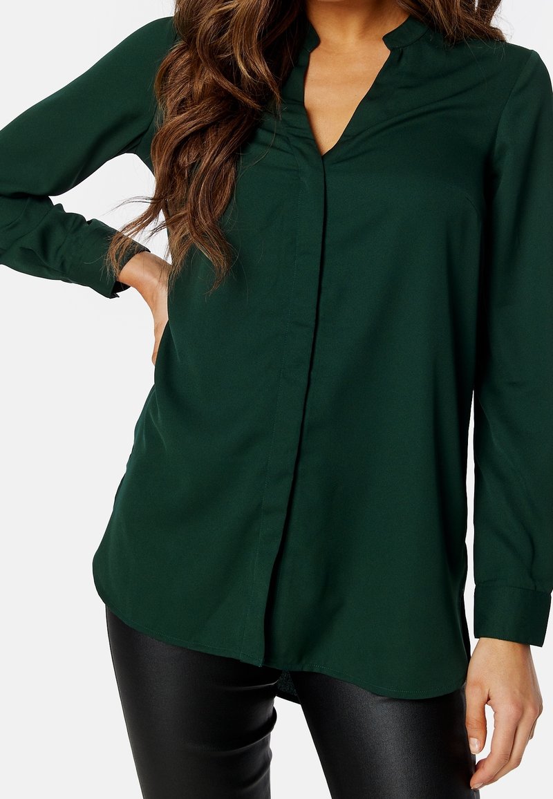 Blouse verte à manches longues avec un décolleté en V, des plis sur le devant et une coupe décontractée. Fabriquée en tissu lisse, avec un ourlet arrondi et des détails de boutons.
