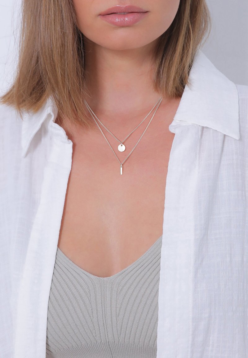 Silver layered necklaces with a circular pendant and a bar pendant, worn over a soft grey ribbed top, framed by a white shirt.