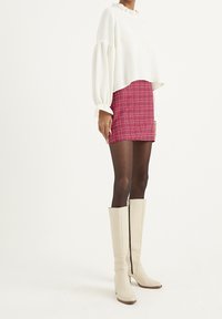 Vit kortärmad blus med volangkrage, kombinerad med en röd rutig kjol. Modellen bär knähöga beige stövlar med sidodragkedja och trädetaljer.