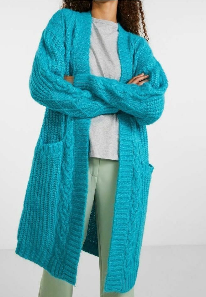 Vrouw met een lange turquoise kabelgebreide cardigan over een grijs T-shirt en lichtgroene broek, met gekruiste armen.