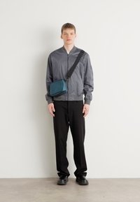 Giacca bomber grigia con zip, pantaloni neri e una borsa a tracolla blu. Materiale liscio, design semplice e vestibilità casual. Scarpe nere.